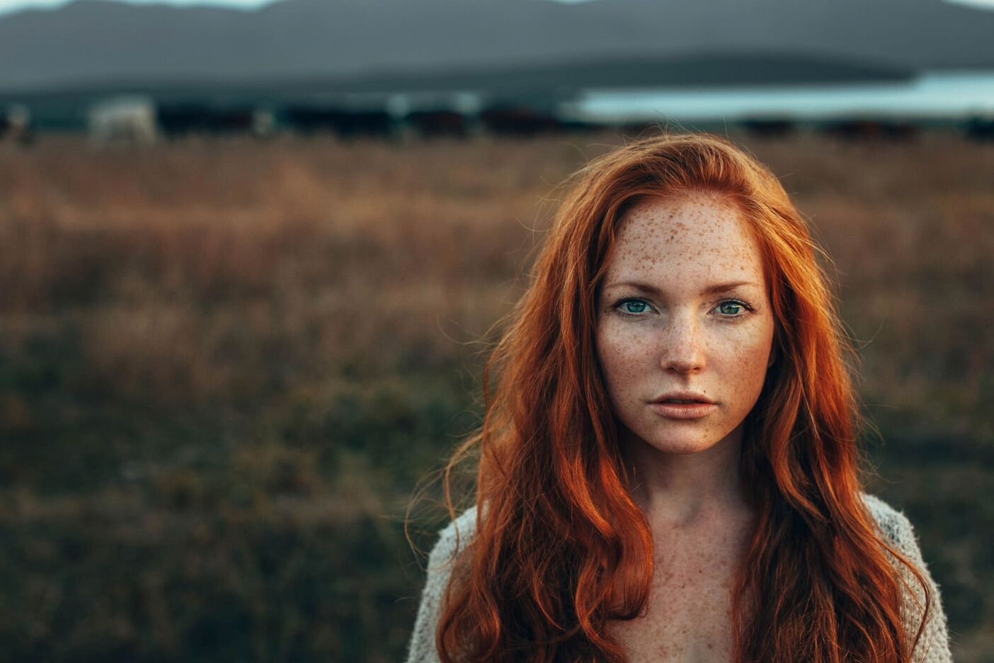 A young woman with long red hair and freckles stands in a grassy field with mountains in the background, reminiscent of a Sunday salon's serene setting. She has a serious expression and is looking directly at the camera.