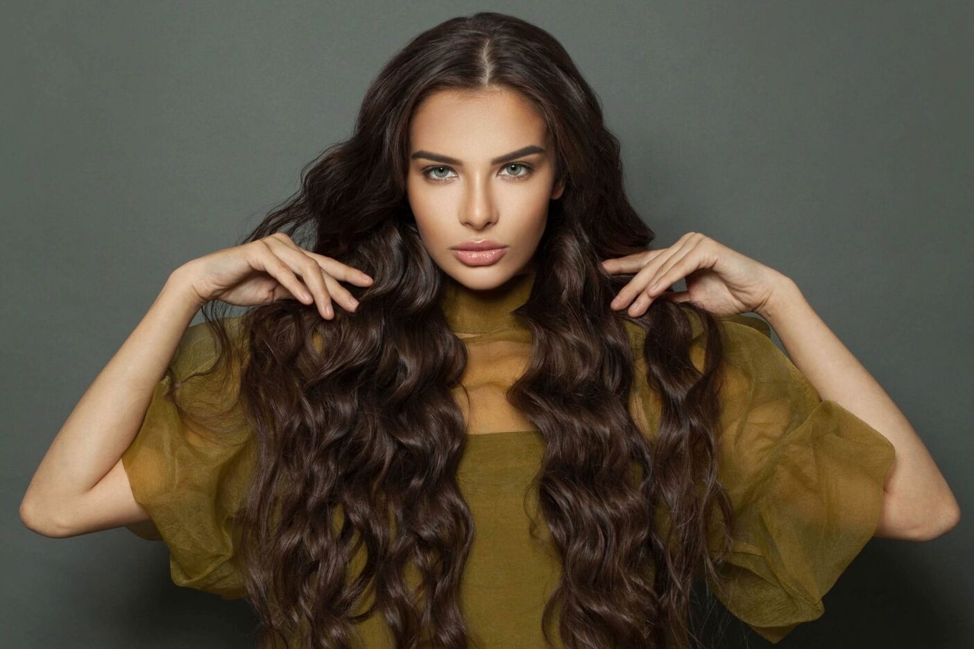 A woman with long, wavy brown hair, wearing a sheer, olive green top, poses against a plain gray background. Her hands are raised to her hair as if showcasing a style from a high-end hair salon near me, and she gazes directly into the camera with a serious expression.