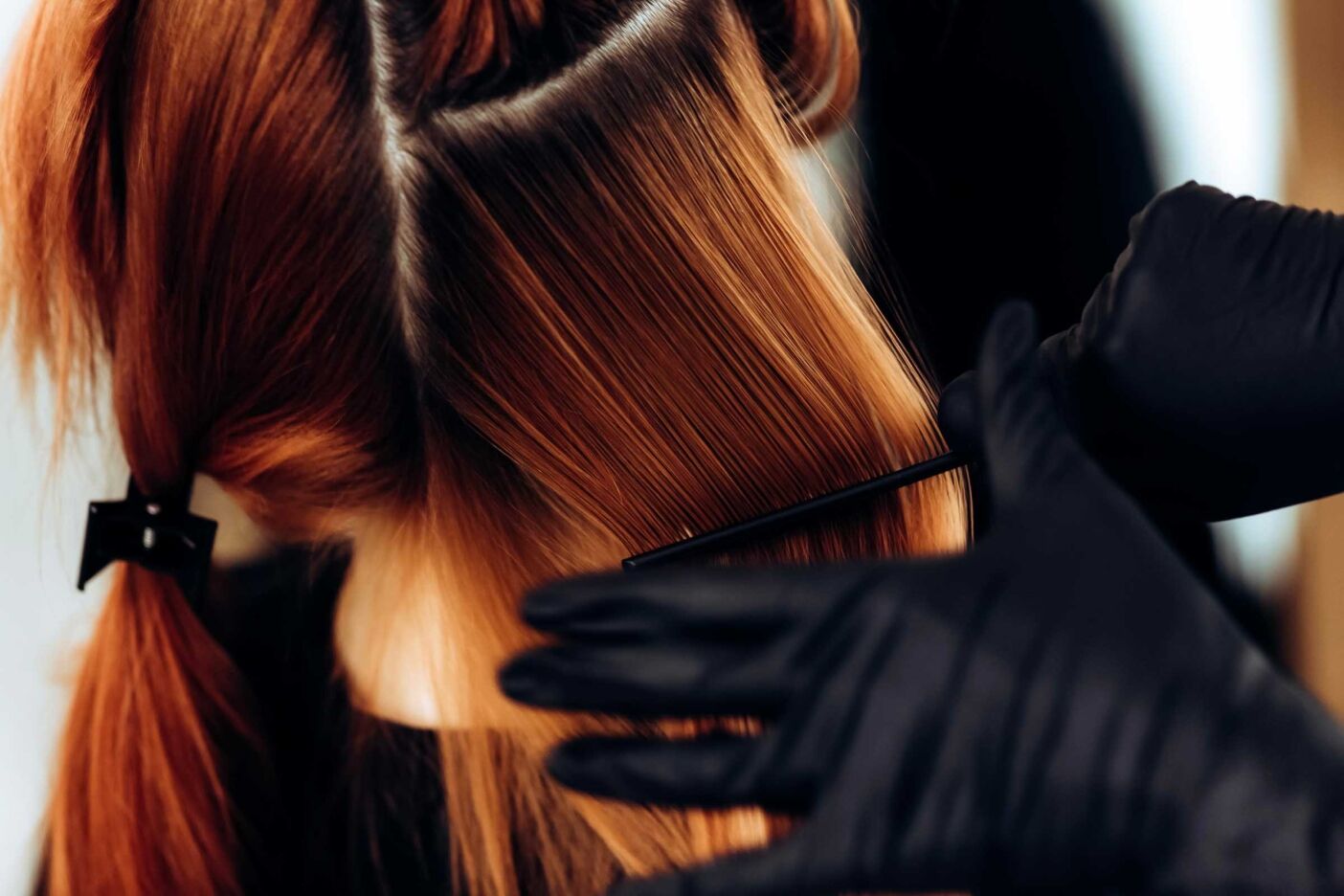 A close-up of a person getting their hair dyed, with a gloved hand applying dye with a comb. The person's hair is sectioned for color corrections, showing a contrast between the natural and dyed hair. The lighting highlights the shine and texture of the hair.