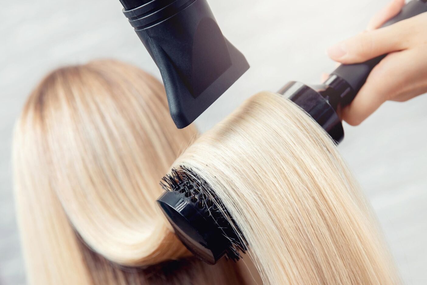 Close-up of a person achieving a blowout in Cary. A round brush shapes and smooths the blonde hair, while a blow dryer with a nozzle attachment targets each section. The setting is bright and well-lit, showcasing the transformation.