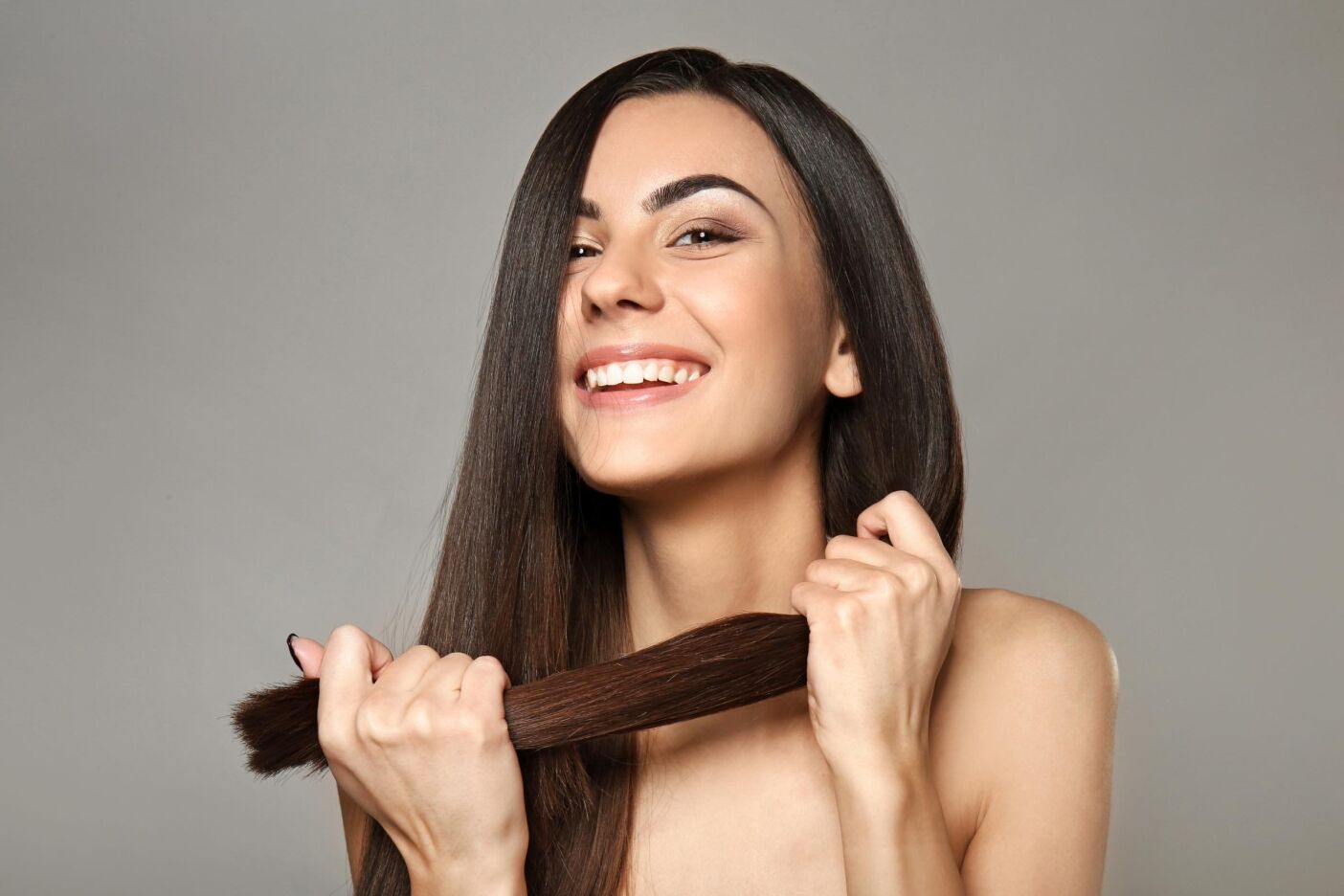 A smiling woman with long, straight brown hair, enhanced by a custom hair treatment, stands against a gray background. She holds her hair with both hands near her chin.