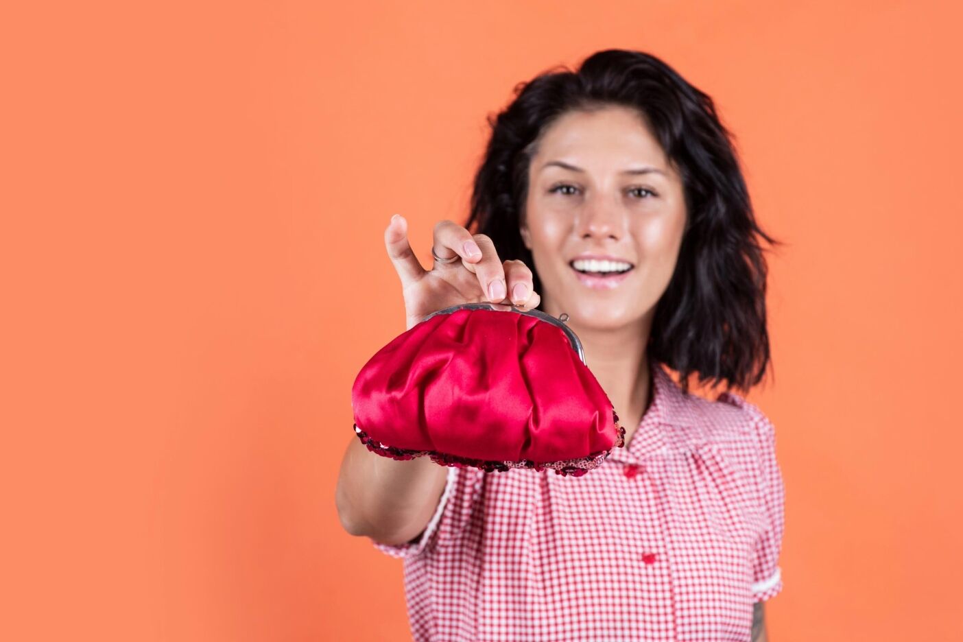 A smiling woman with dark hair and a subtle recession hairline in a red-checkered shirt holds a bright red coin purse toward the camera, standing against an orange background.