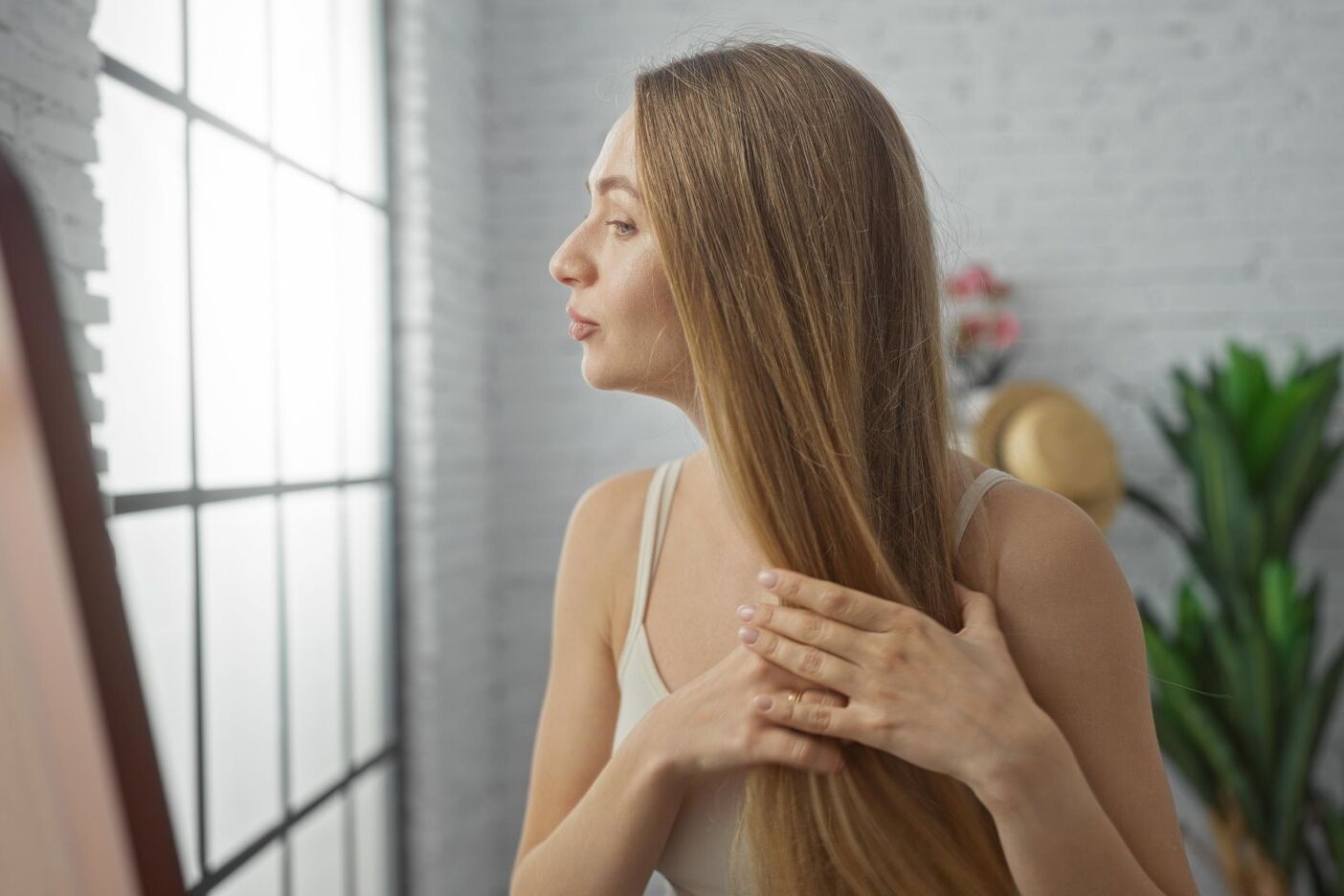 A woman with long, straight brown hair and natural highlights stands indoors, gently touching her hair while looking into a mirror. She wears a light-colored top, and the background features large windows, plants, and a white brick wall.
