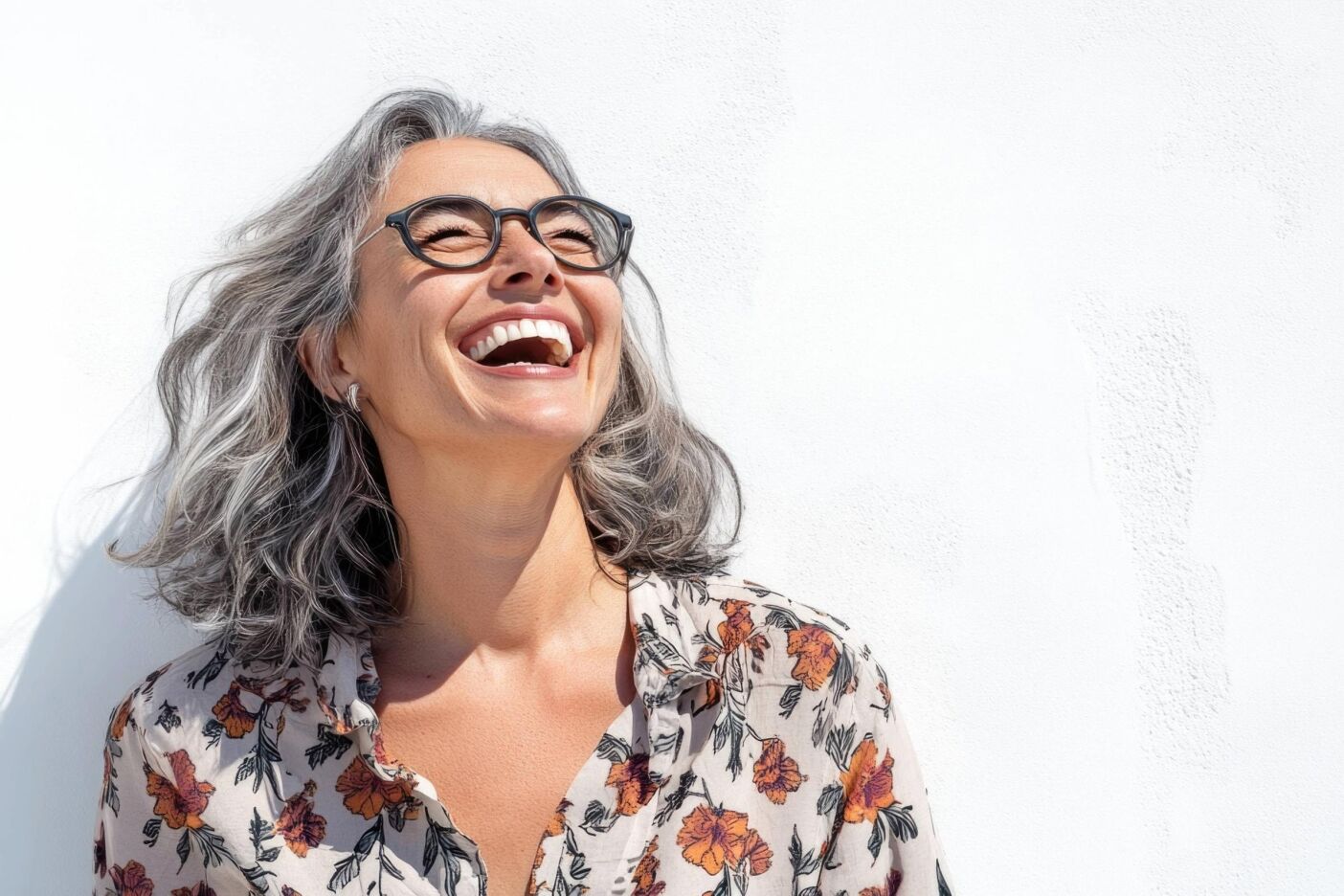 Smiling woman with flawless grey hair and glasses, wearing a floral blouse, stands against a white wall and laughs joyfully, looking upward.